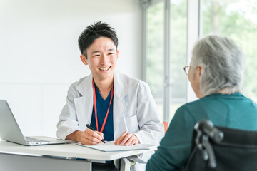 病院・介護施設で高齢者女性を笑顔で診察・問診する若いアジア人男性医師（かかりつけ医）  © buritora