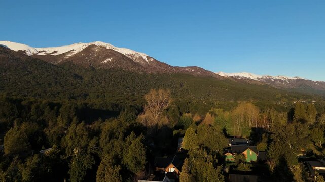 This breathtaking aerial photograph captures the scenic beauty of Cerro Bayo, a mountain located in Villa La Angostura, Argentina. The image showcases a vibrant forested landscape with snow-capped 