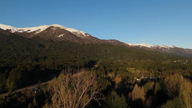 This breathtaking aerial photograph captures the scenic beauty of Cerro Bayo, a mountain located in Villa La Angostura, Argentina. The image showcases a vibrant forested landscape with snow-capped 