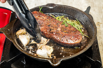 A person is cooking a sirloin steak in a pan with garlic and herbs