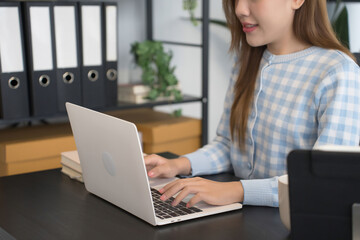 An Asian female businesswoman in a blue-checkered shirt is focused on working on her laptop at a modern office desk, surrounded by neatly organized files and office supplies