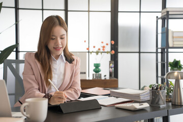 An Asian female businesswoman is seated at her desk, writing notes on a tablet while surrounded by papers and a laptop. The office setting is contemporary and well-organized.