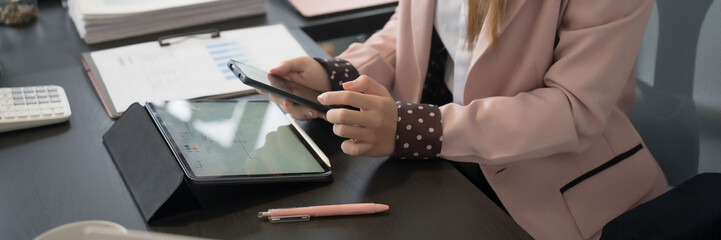 An Asian businesswoman checks her smartphone while working at her desk, surrounded by a laptop, documents, and stationery in a modern office