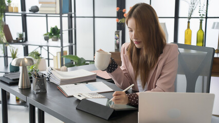 A cheerful Asian businesswoman enjoys a cup of coffee while working at her desk in a bright, modern office. She is engaged in her tasks, with a smile on her face.