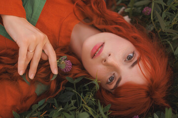 A shepherdess woman with red hair and a red and green dress lees on grass in field of flowers. Rural scene concept with a romantic peasant woman in a linen dress. Romance novel or film.