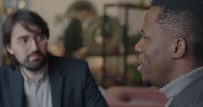 African American businessman talking to Middle Eastern partner and drinking beer from glass during meeting in cafe. Businesspeople and international cooperation concept.