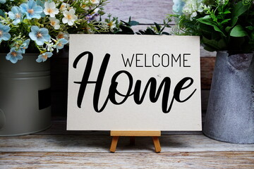 Welcome Home text written on paper card with easel and flowers decoration on wooden background