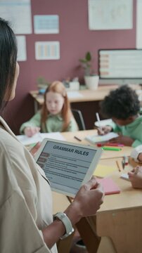Medium vertical over-shoulder shot of female English language teacher with digital tablet explaining grammar rules to class of diverse students at middle school