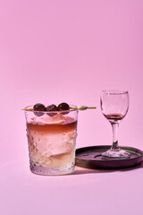 A minimalist still life of two cocktails on a pink background. A clear glass tumbler filled with a light-colored cocktail sits beside a stemmed glass containing a deep red cocktail.