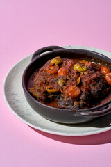 A hearty portion of braised beef cheeks in a rich tomato sauce. The dish is served in a cast iron skillet and garnished with fresh herbs.