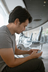 Fototapeta premium Vertical photo of asian Thai man sitting at gym and using mobile phone after done or between resting exercise, workout with dumbbells.