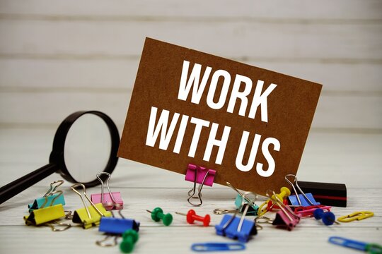 Work With Us text on paper card with magnifying glass and stationary on wooden background