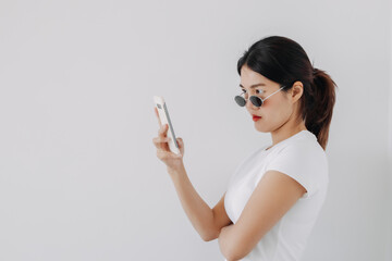 Beautiful and chic asian Thai woman wear vintage sunglasses while using and looking at mobile phone, standing isolated over white background.