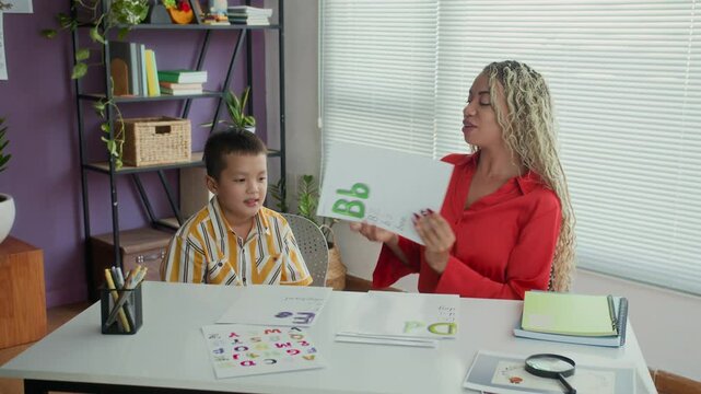 Young female logopedist with dreadlocks training letters pronunciation with Asian teenage kid during lesson at classroom