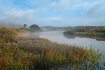 View of an ancient wooden windmill on the banks of the Sorot River on a foggy summer morning, Mikhailovskoye, Pushkinskiye Gory, Pskov region, Russia