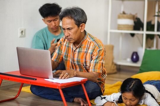 Busy Asian Family Father Working On Laptop, Daughter Drawing While Lying Prone On Floor And Son Learning On Smartpone - Powered by Adobe