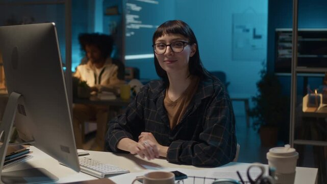Portrait of young female Caucasian database administrator sitting at desk in office of IT companyworking under deadline at nightime, smiling and looking at camera