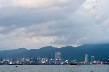 A cloudy sky looms over Shenzhen's bustling port with cranes, ships, and mountains in the...