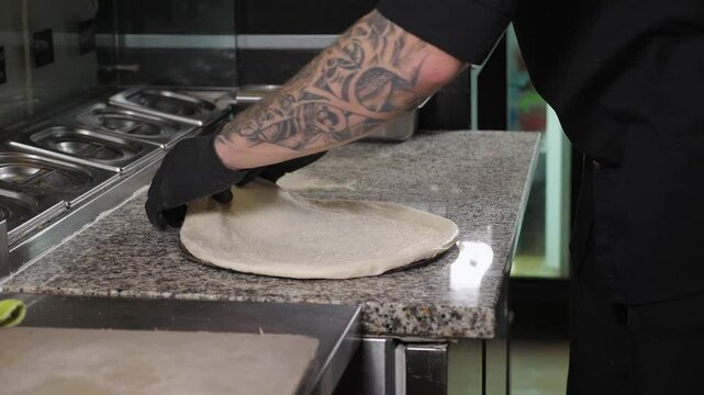 A close-up of a chef in gloves laying out pizza dough on a round baking dish in the restaurant kitchen, the pizza cooking process.