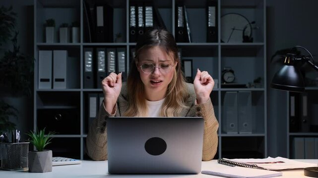 Anxious Caucasian young adult woman waiting deal with crossed fingers while working with laptop in the late night office. Strong desire concept.