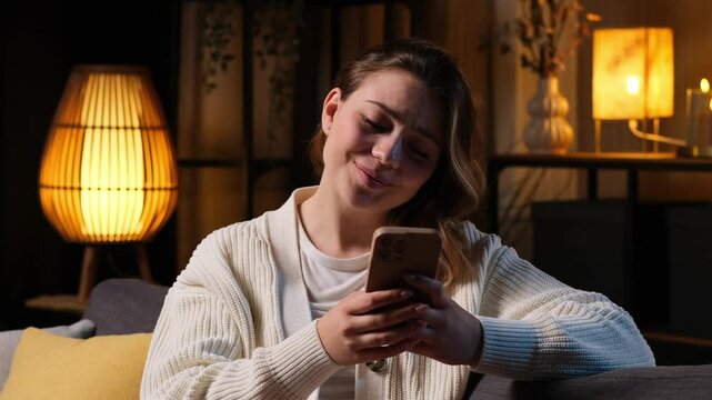 Cheerful Caucasian young woman using phone for texting in online social media with boyfriend at evening home. Spending leisure and free time.