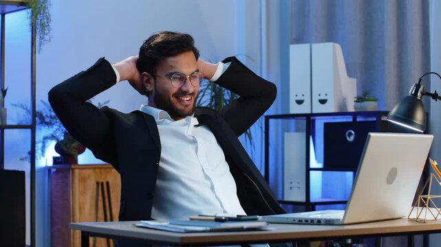 Indian businessman freelancer taking break leaning on chair after working celebrating success victory at modern home office workplace desk. Male Hindu in formal suit puts hands behind head relaxing