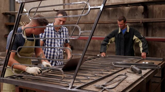 The process of working in the forge, two blacksmiths make metal railings for the stairs, and a customer stands next to him with a laptop and looks at the order options.