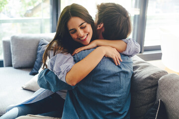 Young millennial couple joyfully embracing on their sofa at home, celebrating a heartfelt proposal involving plans for marriage and starting a family. Their excited expressions capture their happiness