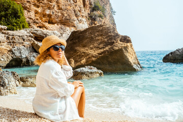 One female tourist sitting at the beach enjoying summer holiday vacation alone wearing straw hat and looking the sun. Blue transparent tropical se ocean water in background. Travel destination