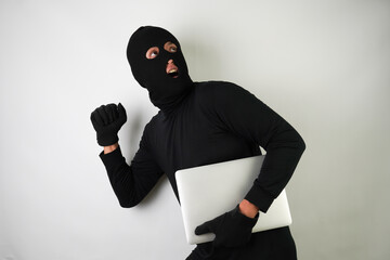 Young Adult black masked robber is holding silver colored laptop. Running, fleeing and escaping pose. Concept for hacking, scamming, phishing.