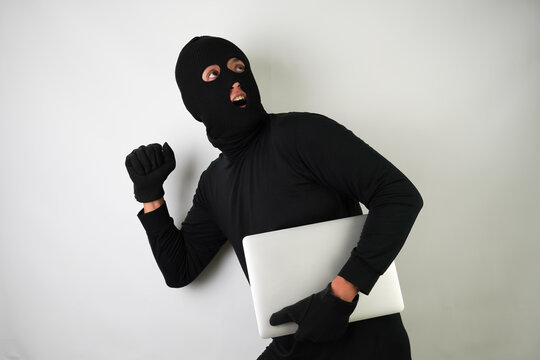 Young Adult black masked robber is holding silver colored laptop. Running, fleeing and escaping pose. Concept for hacking, scamming, phishing.