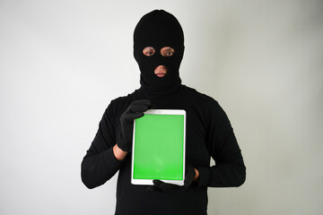 Young Adult black masked robber is holding digital tablet with vertical blank green screen. Concept for hacking, scamming, phishing.