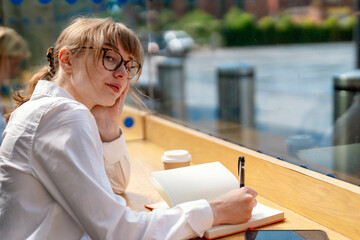 Young Woman in Glasses Thoughtfully Journaling at Cozy Café on Sunny Afternoon