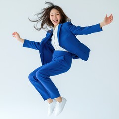 An Asian woman in a business blue suit is captured mid-air, jumping with joy and energy against an isolated background. Her expression radiates happiness and enthusiasm