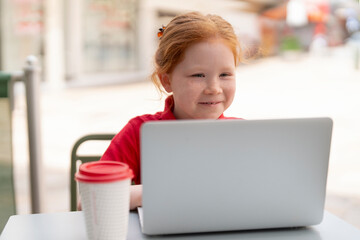 Young Girl Joyfully Working on a Laptop at a Sunny Outdoor Café During a Vibrant Afternoon