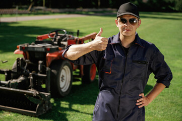 Shows thumb up. Man is with utility tractor with grass cutter and aerator equipment on the field