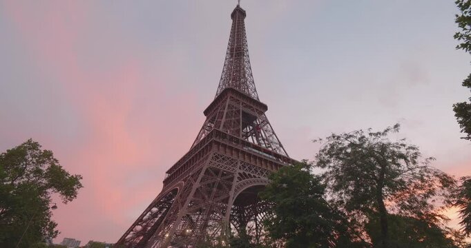 Eiffel Tower during the 2024 Summer Olympics in Paris, France. Early evening pink sky sunset, and night time transition, beautiful day. Cinematic reveal behind tree. Looking up, from below close up.