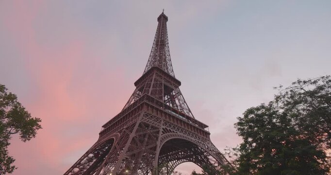 Eiffel Tower during the 2024 Summer Olympics in Paris, France. Early evening sunset, and night time, beautiful pink sky. Cinematic reveal behind tree. Looking up, from below close up.