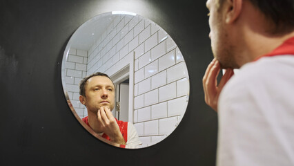 Man in front of mirror smoothes stubble. Men's cosmetics. Young caucasican man preens in front of a mirror.