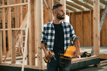 Building the house, holding hard hat and drill. Industrial worker in wooden warehouse