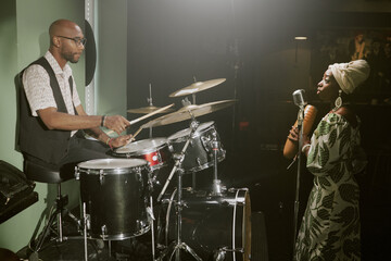 Side view shot of young Black man playing drums and young woman with guiro in hands singing jazz song