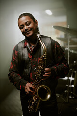 Vertical medium long portrait of joyful African American jazz musician holding saxophone smiling at...