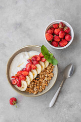 Greek yogurt with muesli, strawberry and banana in a bowl on a light background with fresh berries and spoon.