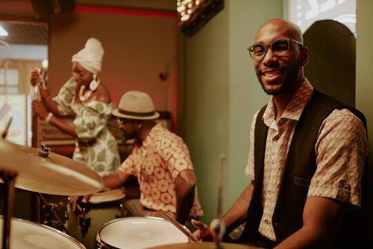 Young African American man sitting at drum kit and smiling at camera while two musicians getting ready for performance in background