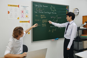 Teacher explaining Pythagorean theorem on chalkboard to student. Both are focused on the lesson, with diagrams and formulas visible on surrounding walls creating an educational atmosphere