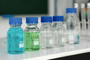 Set of transparent bottles in a laboratory holding different colored liquids, lined up on a white countertop with measuring marks on them and blue screw caps