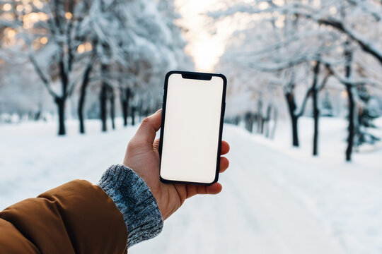 A man's hand holding a smartphone with a blank screen against a wintry background of a snowy park. Horizontal with copy space.