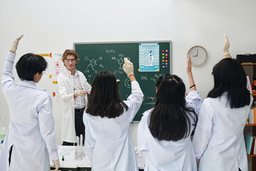 Female instructor wearing lab coat and gloves engaging students with raised hands in classroom while teaching chemistry concepts on chalkboard