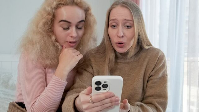 Woman comforting her crying friend showing bad news on mobile phone while sitting on the bed. Upset and distressed girl found out about her boyfriend cheating on social media using smartphone