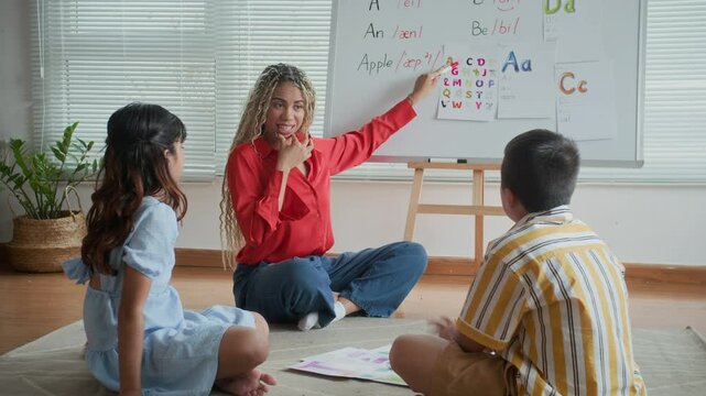 Long shot of young female Latin teacher pointing at whiteboard with educational materials and talking to diverse kids during speech therapy class